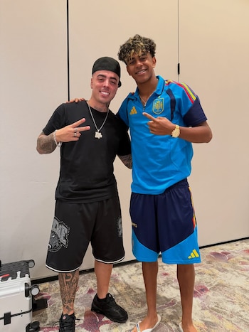 Dos hombres posan de pie en un interior. Uno viste camiseta negra y gorra; el otro, camiseta azul de fútbol con el escudo de España