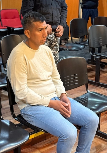 Un hombre con suéter crema y pantalón azul está sentado en una silla negra con las manos juntas en su regazo