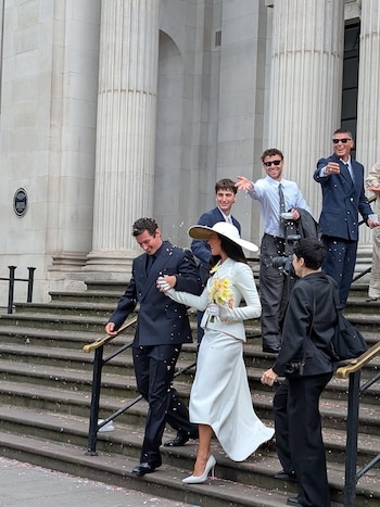 Dua Lipa con vestido blanco y sombrero, de la mano de Callum Turner en traje oscuro, bajan escaleras bajo confeti frente a un edificio con columnas