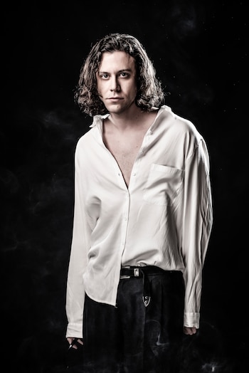 Hombre joven de cabello largo y oscuro, con camisa blanca desabrochada y pantalón oscuro con cinturón, de pie ante un fondo negro con humo