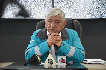 Hombre de cabello canoso con chaqueta azul sentado en mesa oscura con micrófonos, un botón rojo y documentos