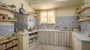 Cocina con paredes de color crema, encimeras de mármol blanco, azulejos decorativos azules y blancos, estantes de madera, ventana con cortina y un fogón.