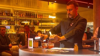 Un bartender sirve un cóctel desde una coctelera a un vaso. Se ven otros vasos, botellas de licor en estantes y varias personas en la barra.
