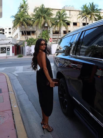 Mujer con vestido negro, gafas y tacones junto a SUV negro en una calle con edificios, palmeras y señales
