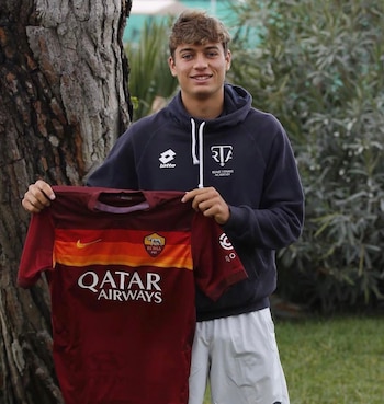 Flavio Cobolli sostiene una camiseta granate y amarilla del AS Roma mientras viste una sudadera oscura en un jardín