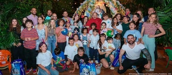 Un grupo grande de hombres, mujeres, niños y niñas posa frente a un arco decorado con luces y una figura religiosa, con plantas y regalos