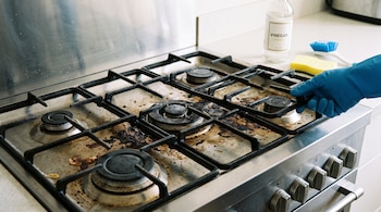 Cocina de gas de acero inoxidable sucia, mano enguantada azul levantando parrilla, botella de vinagre, esponja amarilla, cepillo azul.