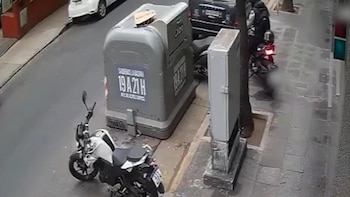Toma de cámara de seguridad que enfoca una calle con una motocicleta blanca, un contenedor gris de basura y la acera