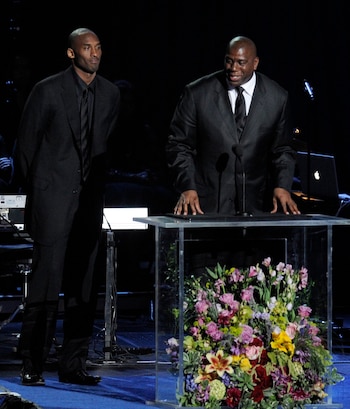 Dos hombres de traje oscuro, Kobe Bryant y Magic Johnson, de pie en un escenario junto a un podio de acrílico transparente con flores