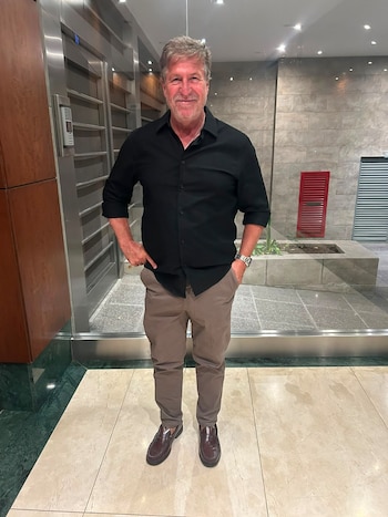 Hombre de pie con camisa negra, pantalón beige y zapatos marrones en un vestíbulo con paredes de piedra y ascensores