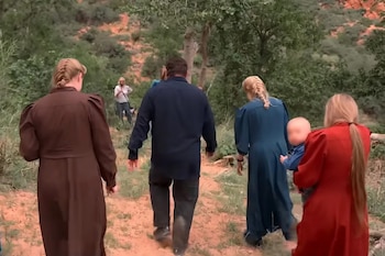 Un grupo de cinco personas, incluyendo un hombre, tres mujeres con vestidos largos y un bebé, camina por un sendero de tierra en un área rural y montañosa