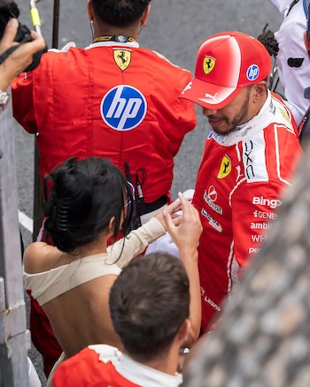 Lewis Hamilton con gorra y traje rojo de Ferrari, siendo abrazado por Kim Kardashian, vista de espaldas con cabello oscuro y top beige