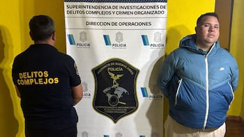 Hombre con chaqueta azul y marcas en el rostro, flanqueado por un oficial de policía de espaldas, frente a un banner de la Policía Bonaerense