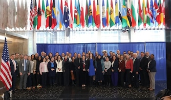 Un gran grupo de personas se reúne frente a un telón azul con banderas de varios países colgando arriba. Una bandera de EE.UU. es visible a la izquierda