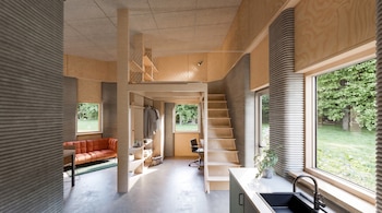 Interior de una vivienda con paredes de textura lineal gris, suelo de cemento, sofá naranja, escritorio, cocina, escaleras de madera y un entrepiso con barandilla.