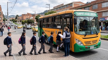 Estudiantes en fila sobre una acera suben a un autobús amarillo y verde junto a una mujer con chaleco identificador y libreta.