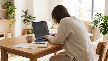 Persona sentada frente a una mesa de madera escribiendo en un portátil junto a un cuaderno, una taza y un teléfono móvil