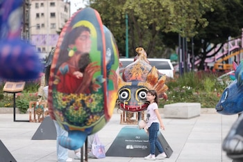 Una niña junto a una escultura multicolor con forma de felino. Otros alebrijes y edificios se aprecian en una plaza urbana