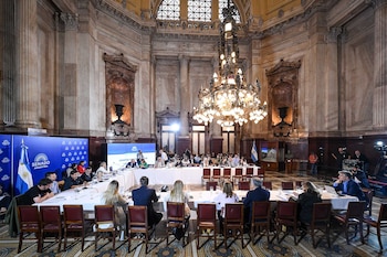 Interior de un gran salón con mesas largas donde personas participan en una reunión. Se aprecian columnas, un candelabro grande y pantallas con el logo del Senado argentino