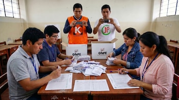 Seis personas en una mesa: cuatro sentadas registrando votos, y dos de pie en el centro grabando con smartphones. Hay cajas blancas con el logo ONPE.