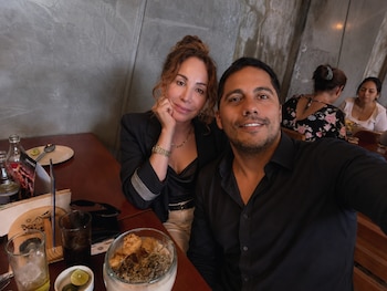 Hombre y mujer sonrientes posan para una fotografía en un restaurante con paredes de concreto, sentados frente a una mesa de madera con comida y bebidas