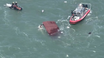 Barco de guardacostas rojo y blanco, moto acuática con dos personas, barco parcialmente hundido con techo rojo, y escombros flotando en el agua
