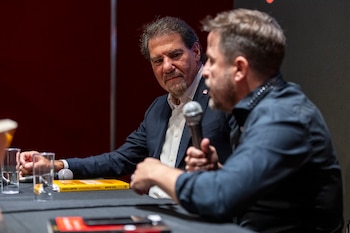 Un hombre de traje oscuro y barba mira a otro hombre barbudo que sostiene un micrófono. Un libro amarillo de economía y vasos de agua están sobre la mesa