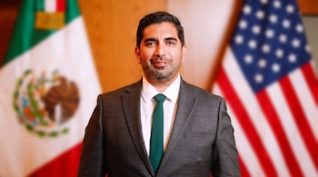 Retrato de un hombre sonriente de tez morena con barba, vestido con traje gris oscuro y corbata verde, frente a las banderas difuminadas de México y Estados Unidos.