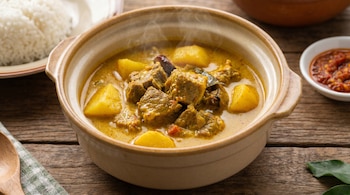 Un tazón de gulai humeante con trozos de carne y patatas en una salsa amarilla cremosa, servido sobre una mesa de madera con arroz y salsa picante.