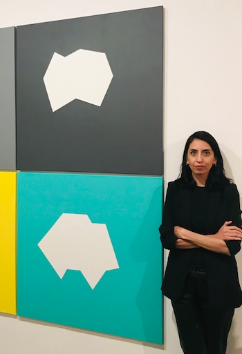 Una mujer de cabello oscuro y chaqueta negra posa junto a una obra de arte modular con formas blancas geométricas sobre fondos grises, turquesas y amarillos