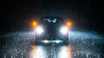 Vista frontal de un coche en la oscuridad bajo una lluvia torrencial, con los faros delanteros y las luces de emergencia naranjas intensamente encendidos.