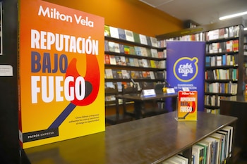 Un ejemplar grande del libro "Reputación Bajo Fuego" de Milton Vela se exhibe en una mesa de madera, con estanterías llenas de libros y un banner azul al fondo