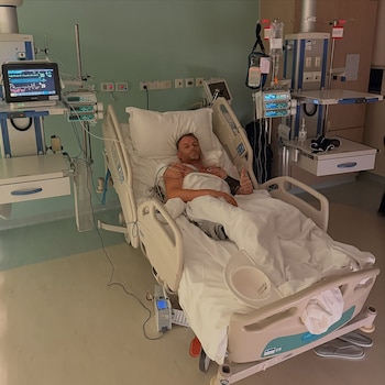 Un hombre en una cama de hospital conectado a varios monitores de control médico levanta el pulgar de su mano izquierda