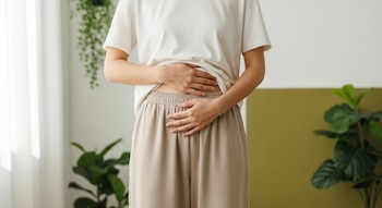 Mujer de pie con camiseta clara y pantalón beige, manos sobre el bajo abdomen. Fondo con plantas verdes y ventana con luz natural.