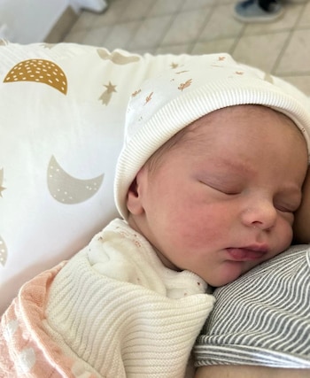 Primer plano de un bebé recién nacido durmiendo con los ojos cerrados, usando un gorro blanco y envuelto en una manta blanca y rosa, apoyado en un hombro a rayas