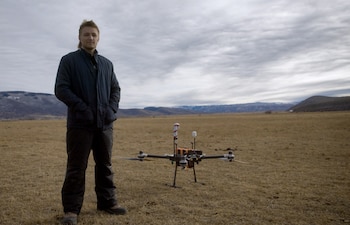 Augustus Doricko, un hombre joven, de pie en un campo seco bajo un cielo nublado, con un dron grande y oscuro a su derecha. Detrás, montañas
