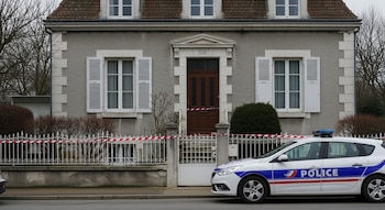 Fachada de vivienda francesa con puerta de madera y ventanas. Un coche de policía francés blanco y azul estacionado. Cinta de acordonamiento roja y blanca en la entrada.