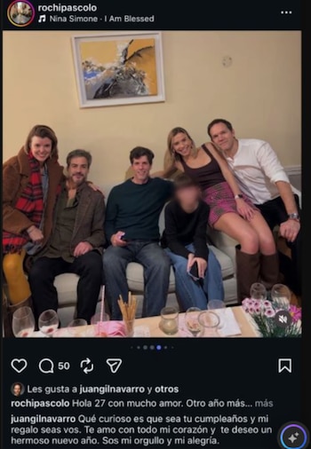 Seis personas sentadas en un sofá y en el suelo frente a una mesa ratona, con copas, comida y un arreglo floral. Hay un cuadro abstracto en la pared