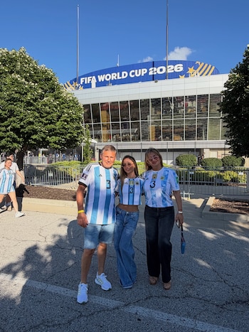 Tres personas adultas posan al aire libre con camisetas de fútbol celestes y blancas. Detrás se observa un estadio con un cartel "FIFA WORLD CUP 2026"