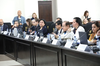 Uma fileira de profissionais sentados em mesas de madeira escura com microfones e jarras de água, alguns olhando para frente e outros conversando entre si