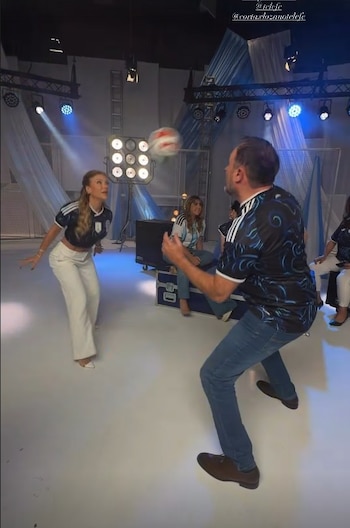 Evangelina Anderson y Augusto Telias juegan con un balón de fútbol en un estudio de televisión. Ella viste una camiseta de fútbol y pantalones blancos, él una camiseta oscura y jeans