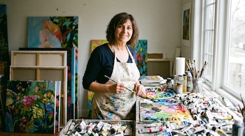 Mujer con delantal manchado de pintura, pincel en mano, sonríe en un estudio. Hay lienzos, tubos de pintura, paletas de colores y pinceles en una mesa.