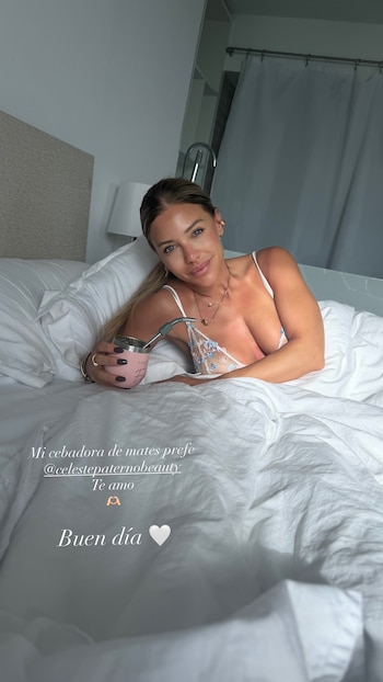 Evangelina Anderson sonríe acostada en una cama blanca, sosteniendo una taza de mate rosa con bombilla, vestida con un top de encaje blanco