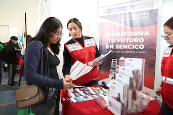Dos mujeres en un stand de Sencico. Una representante con chaleco rojo explica la oferta académica a una joven con gafas. Se aprecian folletos y un banner de Sencico