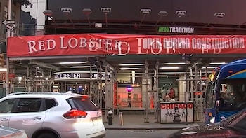 Exterior de Red Lobster en Times Square, con andamios de construcción y un letrero rojo 'RED LOBSTER OPEN DURING CONSTRUCTION'. Coches y un autobús en primer plano