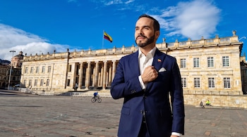 Hombre con traje azul, camisa blanca y pin de bandera colombiana frente al Capitolio Nacional en Bogotá, con bandera de Colombia y cielo azul con nubes.