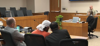 Interior de una sala de tribunal con mesas de madera. Incluye un hombre con turbante blanco, dos hombres con traje, una mujer y un oficial uniformado