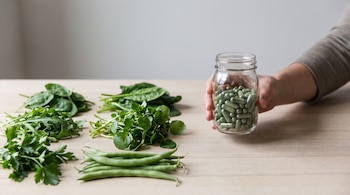 Una mano sostiene un frasco con cápsulas verdes de clorofila. Sobre una mesa, vegetales verdes: espinaca, rúcula, berro, perejil y judías verdes.