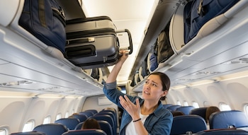 Mujer sostiene una maleta de mano en el pasillo de un avión con compartimentos superiores repletos de equipaje y asientos azules al fondo.