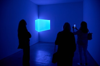 Una sala oscura iluminada por una caja rectangular que emite luz azul brillante en una esquina, con tres siluetas de personas observándola